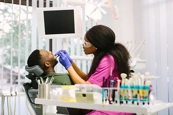 A dentist in a bright clinic examining a male patient's teeth, with dental instruments visible on the desk.